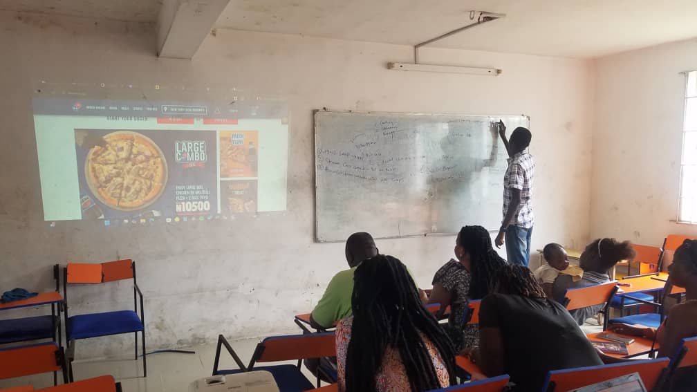 Muyiwa Adekojede writing on the whiteboard beside the Domino's Pizza website projected in class